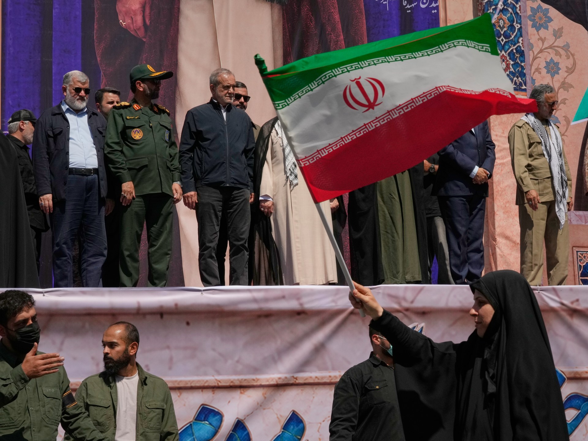 Mohsen Rezaei and other Iranian officials on a public stage behind a large Iranian flag — Al Jazeera liveblog / AP still